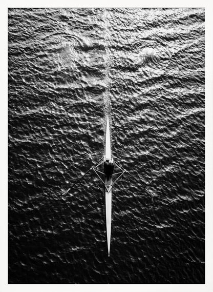 A dramatic black and white overhead photograph of a lone rower in a sleek racing shell, cutting a straight path through dark, textured water. The high-contrast lighting highlights the ripples on the water's surface and the rower's focused form. Artwork