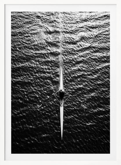 A dramatic black and white overhead photograph of a lone rower in a sleek racing shell, cutting a straight path through dark, textured water. The high-contrast lighting highlights the ripples on the water's surface and the rower's focused form. Artwork