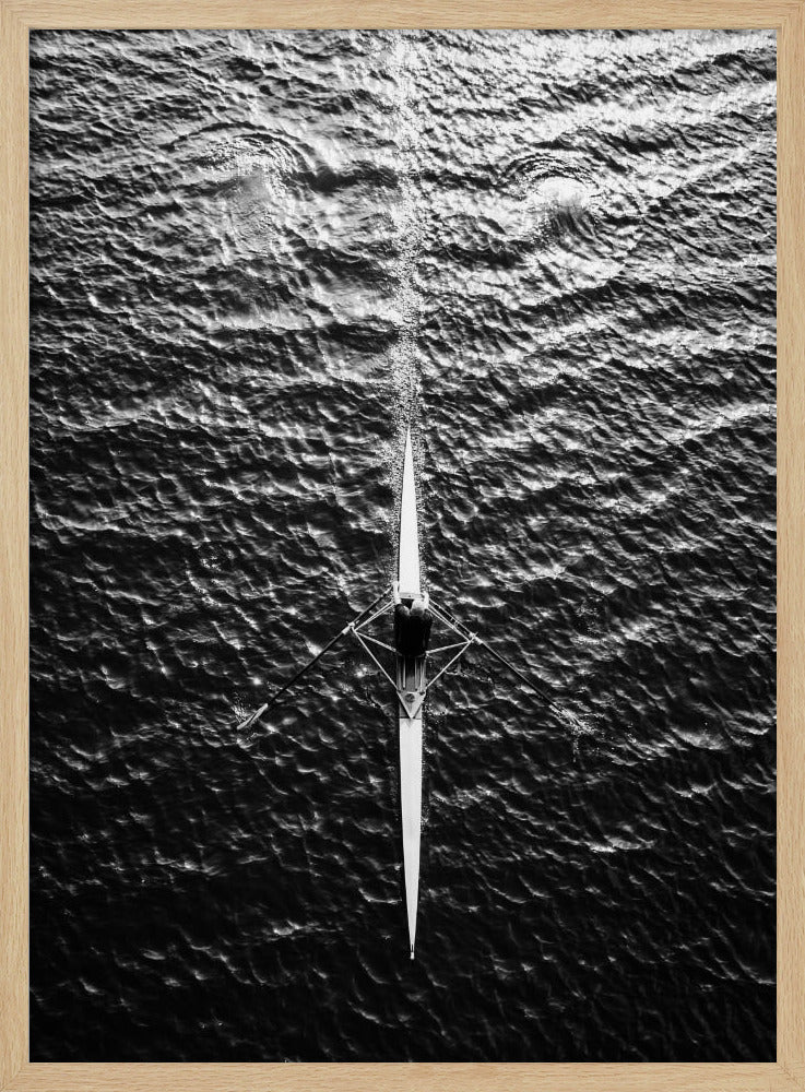 A dramatic black and white overhead photograph of a lone rower in a sleek racing shell, cutting a straight path through dark, textured water. The high-contrast lighting highlights the ripples on the water's surface and the rower's focused form. Artwork