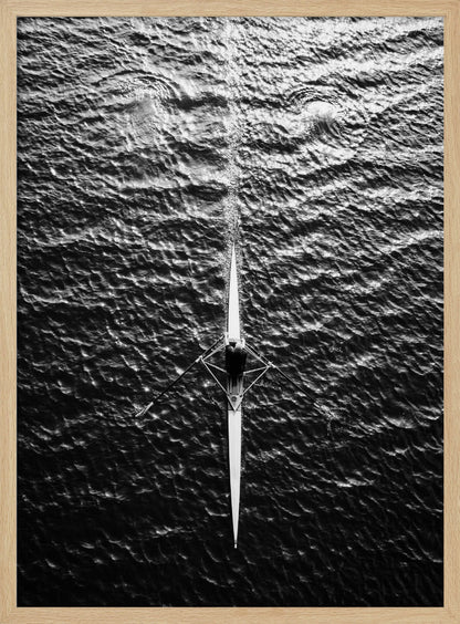A dramatic black and white overhead photograph of a lone rower in a sleek racing shell, cutting a straight path through dark, textured water. The high-contrast lighting highlights the ripples on the water's surface and the rower's focused form. Artwork