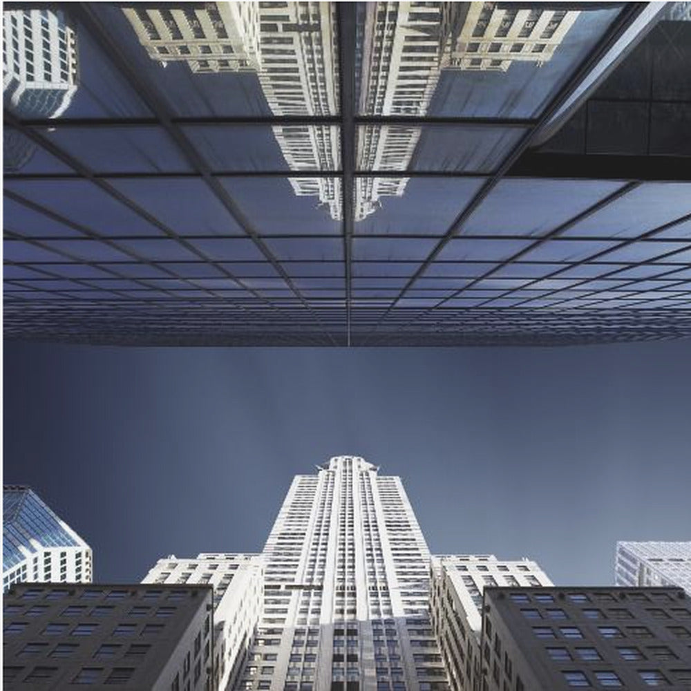 A framed, low-angle photograph capturing a stark white skyscraper against a deep blue sky, with its perfect reflection mirrored in the dark glass facade of an overhanging building, creating a surreal and symmetrical composition. Wall Art
