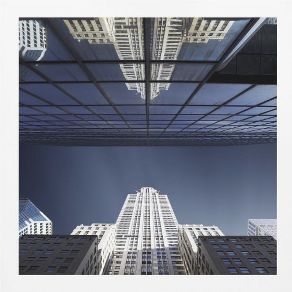 A framed, low-angle photograph capturing a stark white skyscraper against a deep blue sky, with its perfect reflection mirrored in the dark glass facade of an overhanging building, creating a surreal and symmetrical composition. Wall Art