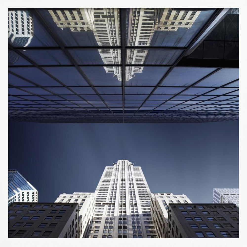 A framed, low-angle photograph capturing a stark white skyscraper against a deep blue sky, with its perfect reflection mirrored in the dark glass facade of an overhanging building, creating a surreal and symmetrical composition. Wall Art