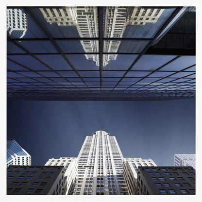A framed, low-angle photograph capturing a stark white skyscraper against a deep blue sky, with its perfect reflection mirrored in the dark glass facade of an overhanging building, creating a surreal and symmetrical composition. Wall Art