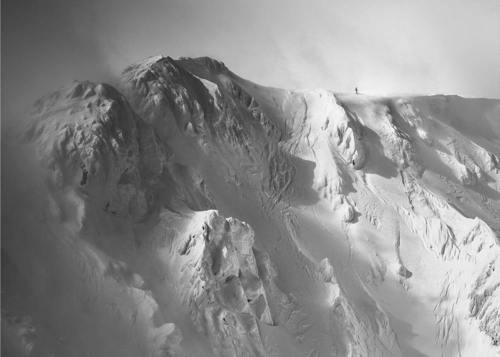 A dramatic black and white photograph of a person standing alone on the ridge of a massive, snow-covered mountain, emphasizing the scale and solitude of the vast, rugged landscape, all presented in a silver frame. Wall Art