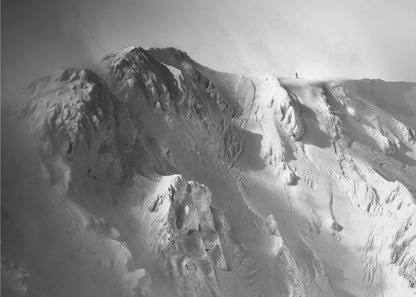 A dramatic black and white photograph of a person standing alone on the ridge of a massive, snow-covered mountain, emphasizing the scale and solitude of the vast, rugged landscape, all presented in a silver frame. Wall Art