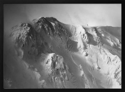 A dramatic black and white photograph of a person standing alone on the ridge of a massive, snow-covered mountain, emphasizing the scale and solitude of the vast, rugged landscape, all presented in a silver frame. Wall Art