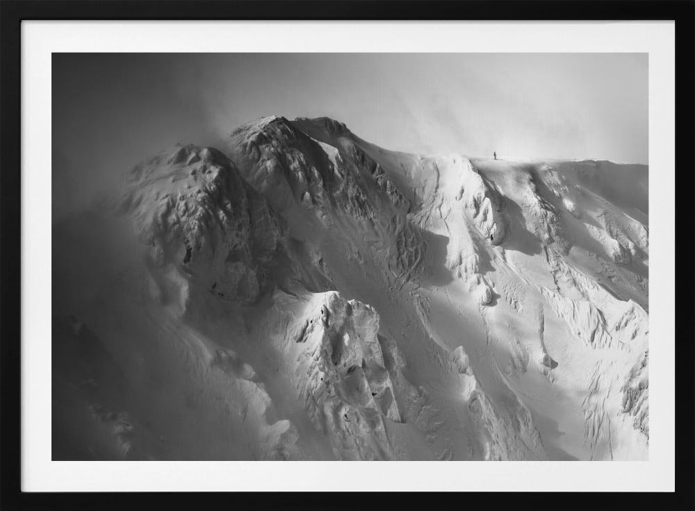 A dramatic black and white photograph of a person standing alone on the ridge of a massive, snow-covered mountain, emphasizing the scale and solitude of the vast, rugged landscape, all presented in a silver frame. Wall Art