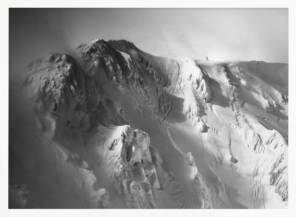 A dramatic black and white photograph of a person standing alone on the ridge of a massive, snow-covered mountain, emphasizing the scale and solitude of the vast, rugged landscape, all presented in a silver frame. Wall Art