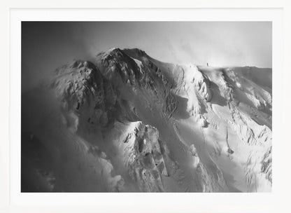 A dramatic black and white photograph of a person standing alone on the ridge of a massive, snow-covered mountain, emphasizing the scale and solitude of the vast, rugged landscape, all presented in a silver frame. Wall Art