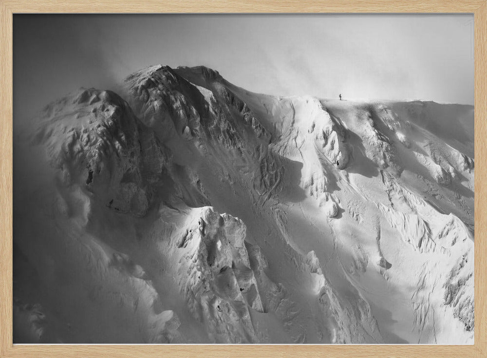A dramatic black and white photograph of a person standing alone on the ridge of a massive, snow-covered mountain, emphasizing the scale and solitude of the vast, rugged landscape, all presented in a silver frame. Wall Art
