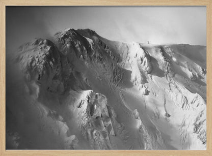 A dramatic black and white photograph of a person standing alone on the ridge of a massive, snow-covered mountain, emphasizing the scale and solitude of the vast, rugged landscape, all presented in a silver frame. Wall Art