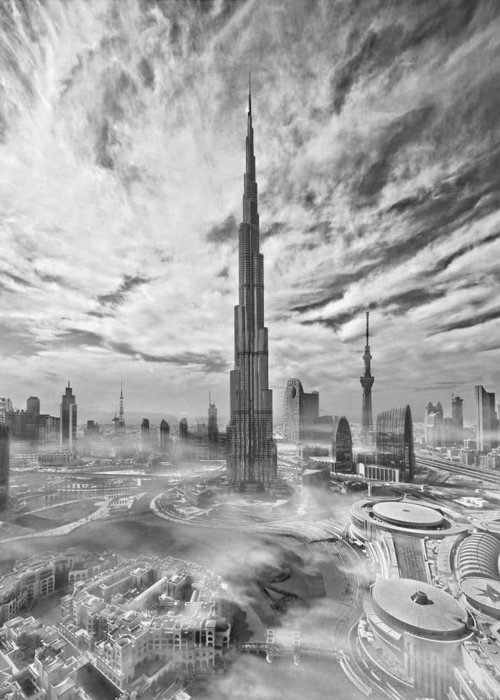 A dramatic black and white high-angle photograph of the Burj Khalifa dominating the Dubai cityscape, with swirling clouds above and a light mist surrounding the buildings below, all enclosed in a black frame. Poster