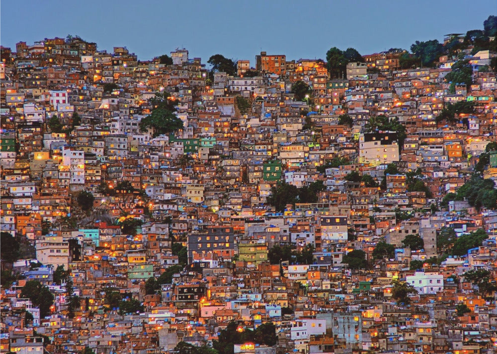 A photograph of a densely packed favela built on a hillside at dusk. The numerous small, colorful houses are illuminated from within, casting a warm orange glow that contrasts with the deep blue of the twilight sky. The image is presented within a silver picture frame. Artwork