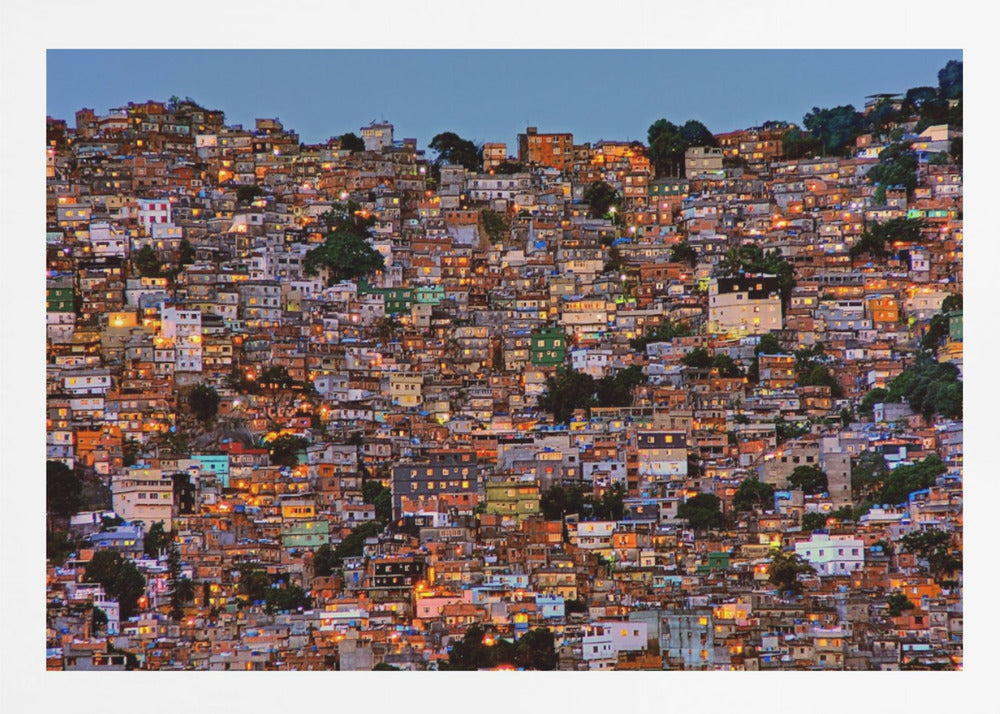 A photograph of a densely packed favela built on a hillside at dusk. The numerous small, colorful houses are illuminated from within, casting a warm orange glow that contrasts with the deep blue of the twilight sky. The image is presented within a silver picture frame. Artwork