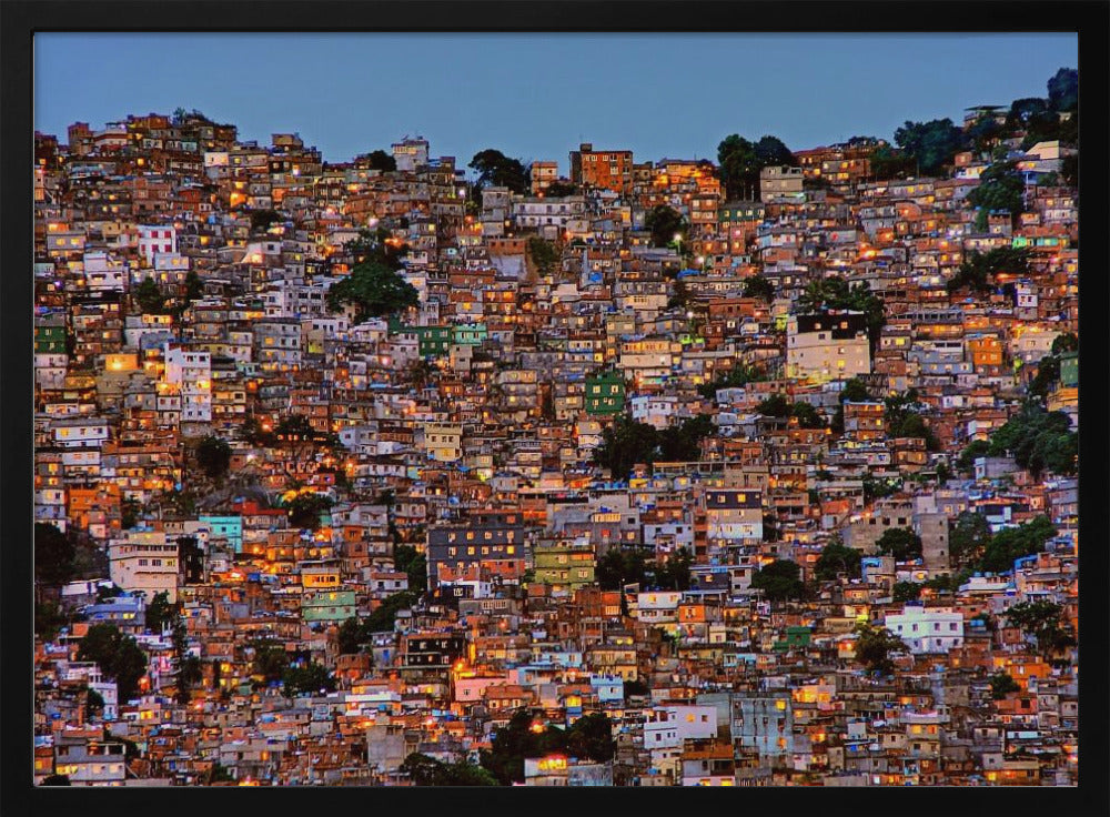 A photograph of a densely packed favela built on a hillside at dusk. The numerous small, colorful houses are illuminated from within, casting a warm orange glow that contrasts with the deep blue of the twilight sky. The image is presented within a silver picture frame. Artwork