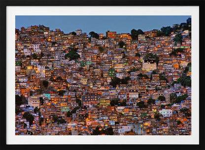 A photograph of a densely packed favela built on a hillside at dusk. The numerous small, colorful houses are illuminated from within, casting a warm orange glow that contrasts with the deep blue of the twilight sky. The image is presented within a silver picture frame. Artwork