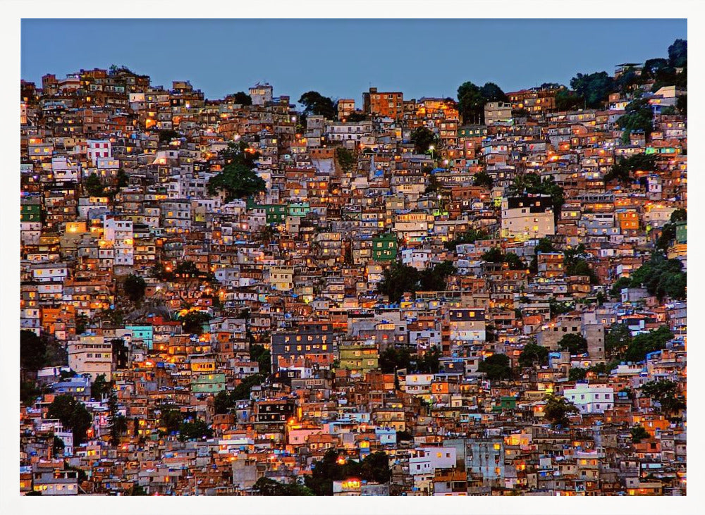A photograph of a densely packed favela built on a hillside at dusk. The numerous small, colorful houses are illuminated from within, casting a warm orange glow that contrasts with the deep blue of the twilight sky. The image is presented within a silver picture frame. Artwork