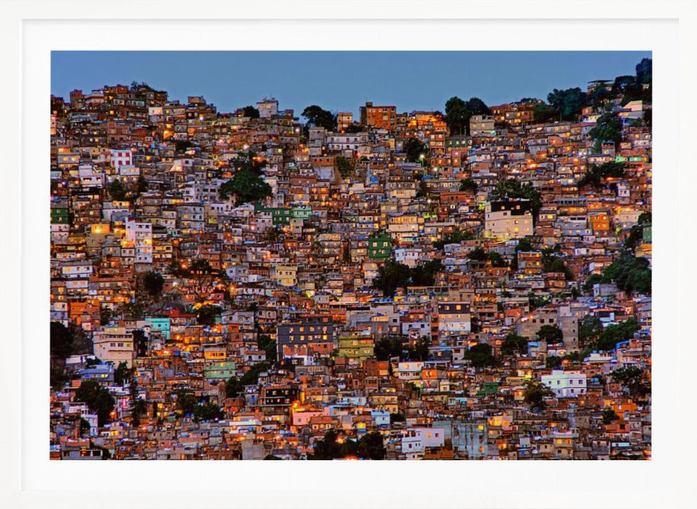 A photograph of a densely packed favela built on a hillside at dusk. The numerous small, colorful houses are illuminated from within, casting a warm orange glow that contrasts with the deep blue of the twilight sky. The image is presented within a silver picture frame. Artwork