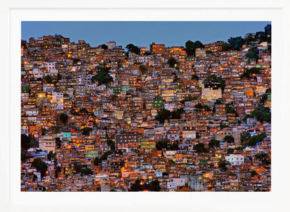A photograph of a densely packed favela built on a hillside at dusk. The numerous small, colorful houses are illuminated from within, casting a warm orange glow that contrasts with the deep blue of the twilight sky. The image is presented within a silver picture frame. Artwork