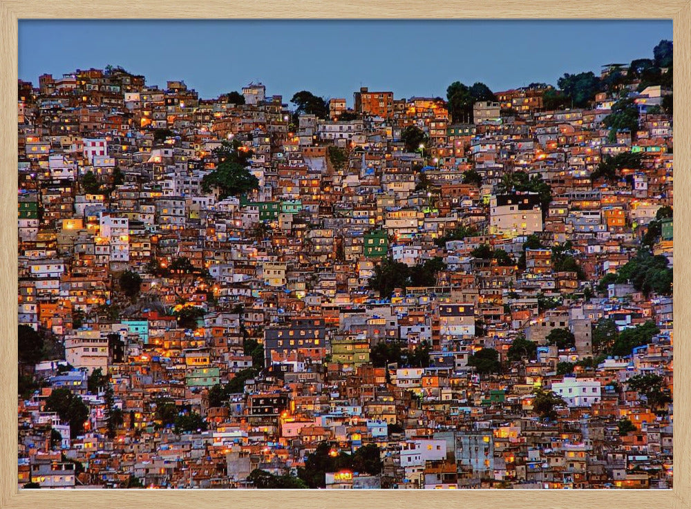 A photograph of a densely packed favela built on a hillside at dusk. The numerous small, colorful houses are illuminated from within, casting a warm orange glow that contrasts with the deep blue of the twilight sky. The image is presented within a silver picture frame. Artwork
