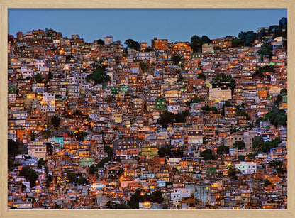 A photograph of a densely packed favela built on a hillside at dusk. The numerous small, colorful houses are illuminated from within, casting a warm orange glow that contrasts with the deep blue of the twilight sky. The image is presented within a silver picture frame. Artwork