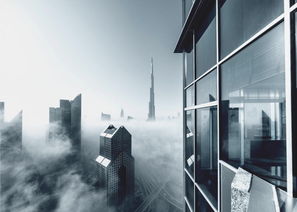 A framed black and white photograph with a cool blue tint, depicting the Dubai skyline from a high-rise window, with skyscrapers including the Burj Khalifa piercing through a thick blanket of fog. Artwork