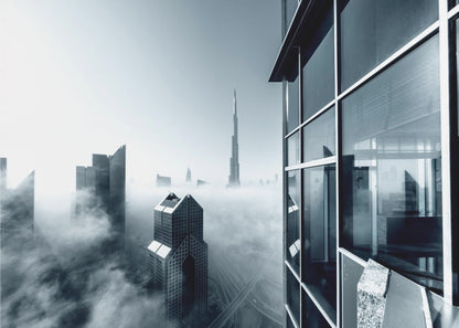 A framed black and white photograph with a cool blue tint, depicting the Dubai skyline from a high-rise window, with skyscrapers including the Burj Khalifa piercing through a thick blanket of fog. Artwork