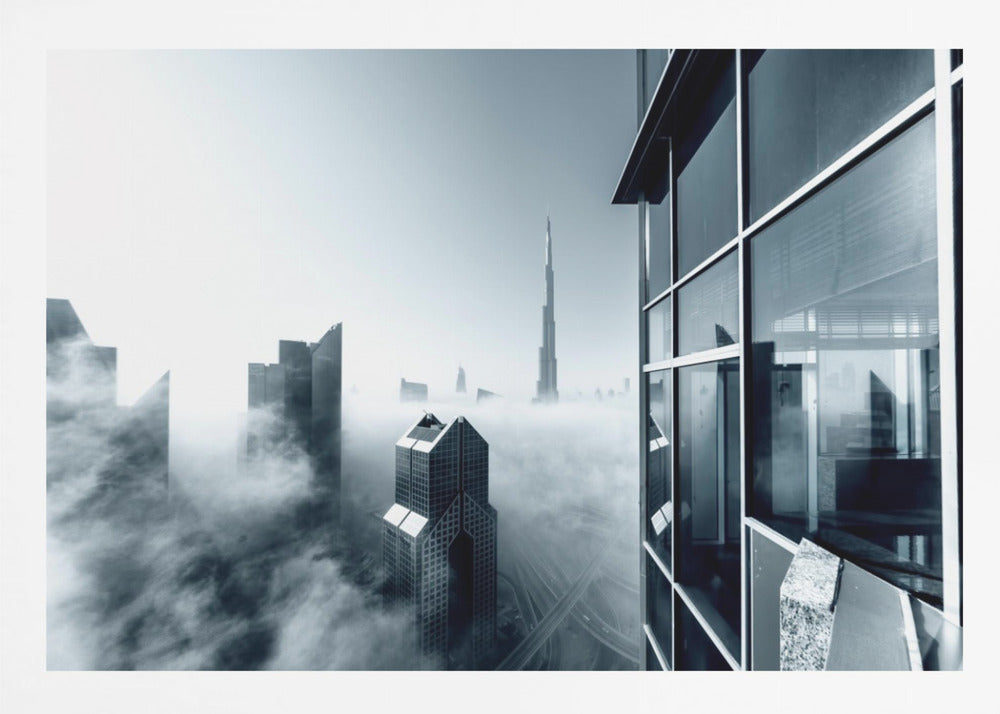 A framed black and white photograph with a cool blue tint, depicting the Dubai skyline from a high-rise window, with skyscrapers including the Burj Khalifa piercing through a thick blanket of fog. Artwork