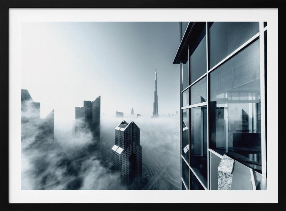 A framed black and white photograph with a cool blue tint, depicting the Dubai skyline from a high-rise window, with skyscrapers including the Burj Khalifa piercing through a thick blanket of fog. Artwork