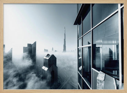 A framed black and white photograph with a cool blue tint, depicting the Dubai skyline from a high-rise window, with skyscrapers including the Burj Khalifa piercing through a thick blanket of fog. Artwork