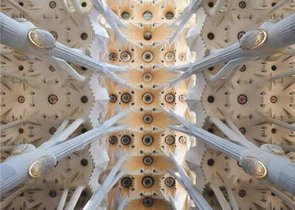 A symmetrical photograph looking directly up at the ornate, geometric ceiling of the Sagrada Familia. White, tree-like columns branch out to support the intricate vaulted ceiling, which is detailed with warm orange and gold starburst patterns. Decor