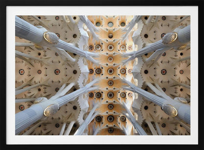 A symmetrical photograph looking directly up at the ornate, geometric ceiling of the Sagrada Familia. White, tree-like columns branch out to support the intricate vaulted ceiling, which is detailed with warm orange and gold starburst patterns. Decor