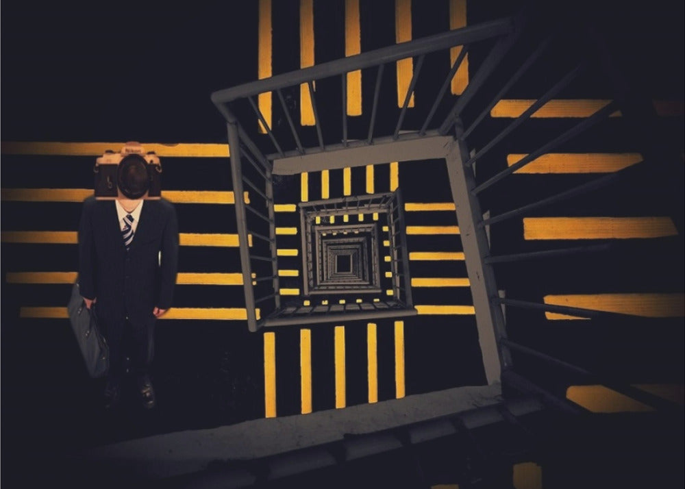 A surreal high-angle view looking down an infinite square spiral staircase with black steps and bold yellow stripes. On a landing stands a man in a dark suit and tie, holding a briefcase, with a vintage camera in place of his head. Artwork