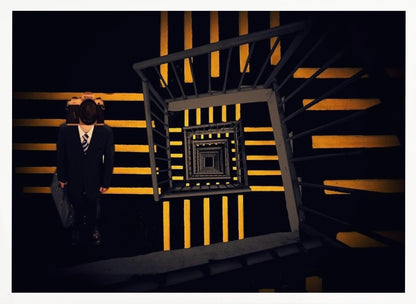 A surreal high-angle view looking down an infinite square spiral staircase with black steps and bold yellow stripes. On a landing stands a man in a dark suit and tie, holding a briefcase, with a vintage camera in place of his head. Artwork