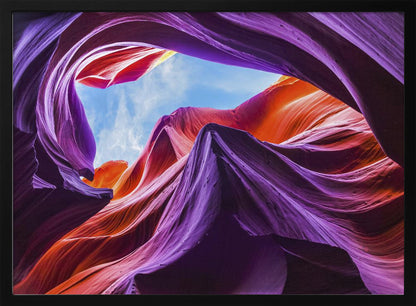 A low-angle photograph looking up from inside a vibrant slot canyon. The smooth, flowing sandstone walls are colored in rich shades of purple, red, and orange, curving around an opening that reveals a bright blue sky with wispy clouds. The image is presented within a silver-colored frame. Wall Art