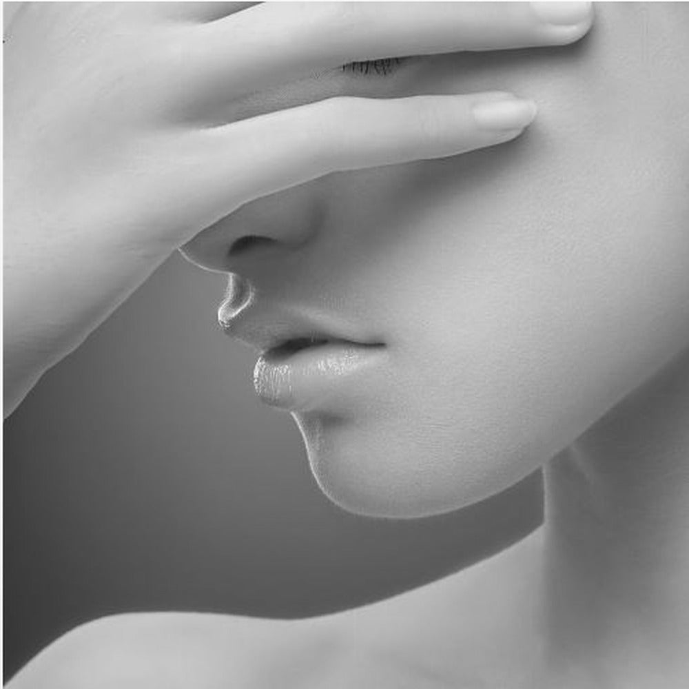 A close-up, black and white photograph of a woman's face. Her hand covers her eyes, revealing only the lower half of her face including her nose, lips, and chin. The image has a soft, sensual quality and is set within a light-colored wooden frame. Decor