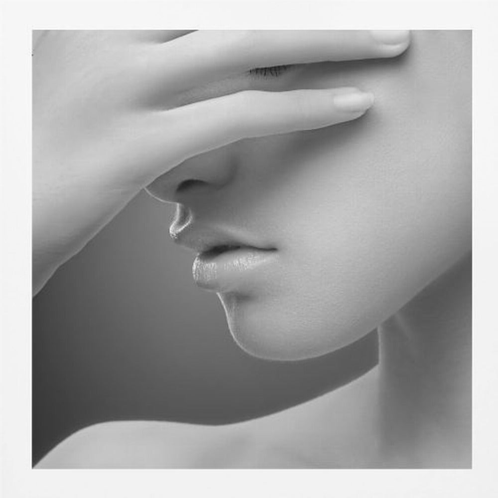 A close-up, black and white photograph of a woman's face. Her hand covers her eyes, revealing only the lower half of her face including her nose, lips, and chin. The image has a soft, sensual quality and is set within a light-colored wooden frame. Decor