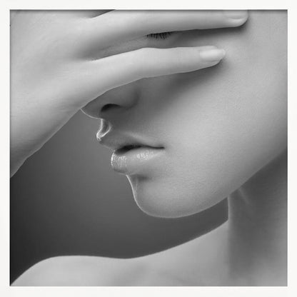 A close-up, black and white photograph of a woman's face. Her hand covers her eyes, revealing only the lower half of her face including her nose, lips, and chin. The image has a soft, sensual quality and is set within a light-colored wooden frame. Decor