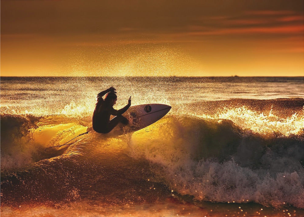 A silhouetted surfer carves a turn on a sun-kissed wave during a vibrant orange sunset, with golden light reflecting off the water and sea spray. Wall Art