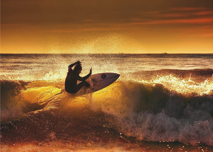 A silhouetted surfer carves a turn on a sun-kissed wave during a vibrant orange sunset, with golden light reflecting off the water and sea spray. Wall Art