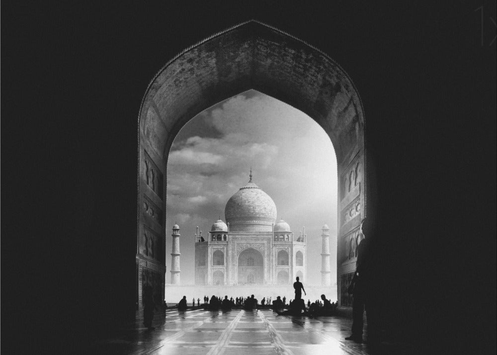 A dramatic black and white photograph of the Taj Mahal seen through a large, dark archway which frames the view. The iconic mausoleum is in the distance under a cloudy sky, and the silhouettes of many people are scattered on the wet, reflective ground in the foreground. Decor
