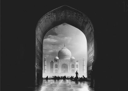 A dramatic black and white photograph of the Taj Mahal seen through a large, dark archway which frames the view. The iconic mausoleum is in the distance under a cloudy sky, and the silhouettes of many people are scattered on the wet, reflective ground in the foreground. Decor