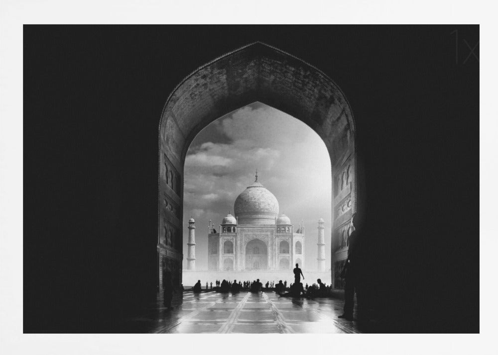 A dramatic black and white photograph of the Taj Mahal seen through a large, dark archway which frames the view. The iconic mausoleum is in the distance under a cloudy sky, and the silhouettes of many people are scattered on the wet, reflective ground in the foreground. Decor