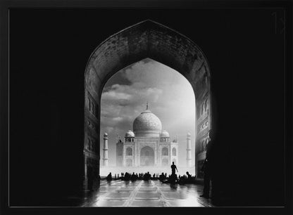 A dramatic black and white photograph of the Taj Mahal seen through a large, dark archway which frames the view. The iconic mausoleum is in the distance under a cloudy sky, and the silhouettes of many people are scattered on the wet, reflective ground in the foreground. Decor