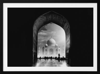A dramatic black and white photograph of the Taj Mahal seen through a large, dark archway which frames the view. The iconic mausoleum is in the distance under a cloudy sky, and the silhouettes of many people are scattered on the wet, reflective ground in the foreground. Decor