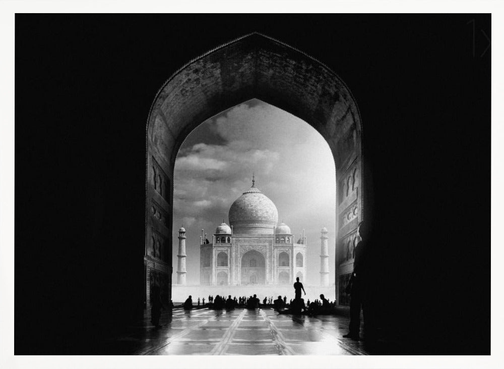 A dramatic black and white photograph of the Taj Mahal seen through a large, dark archway which frames the view. The iconic mausoleum is in the distance under a cloudy sky, and the silhouettes of many people are scattered on the wet, reflective ground in the foreground. Decor