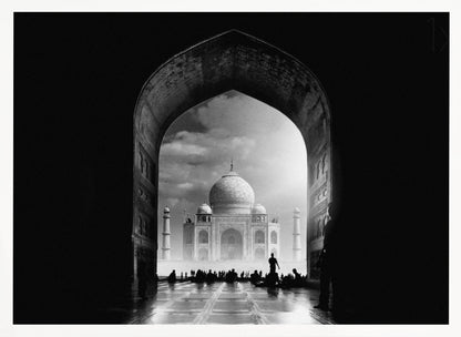 A dramatic black and white photograph of the Taj Mahal seen through a large, dark archway which frames the view. The iconic mausoleum is in the distance under a cloudy sky, and the silhouettes of many people are scattered on the wet, reflective ground in the foreground. Decor
