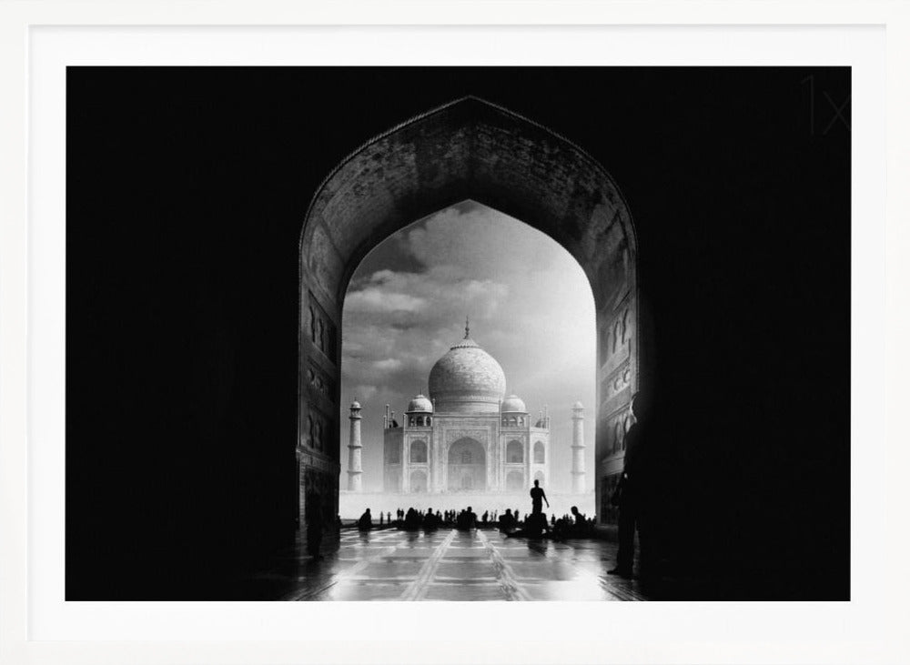 A dramatic black and white photograph of the Taj Mahal seen through a large, dark archway which frames the view. The iconic mausoleum is in the distance under a cloudy sky, and the silhouettes of many people are scattered on the wet, reflective ground in the foreground. Decor