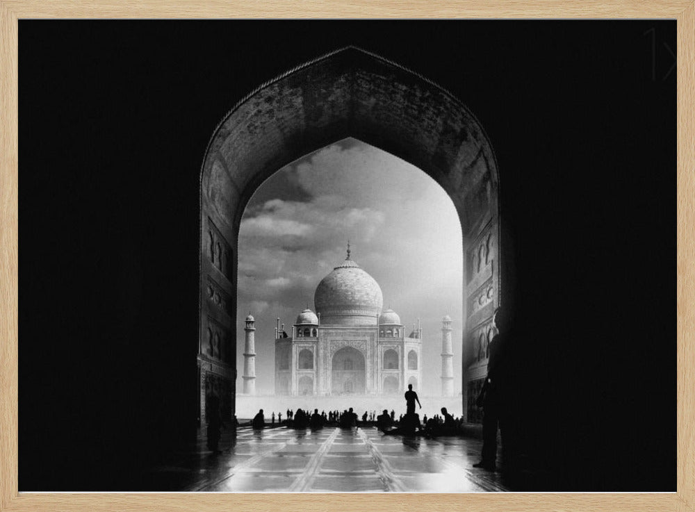 A dramatic black and white photograph of the Taj Mahal seen through a large, dark archway which frames the view. The iconic mausoleum is in the distance under a cloudy sky, and the silhouettes of many people are scattered on the wet, reflective ground in the foreground. Decor